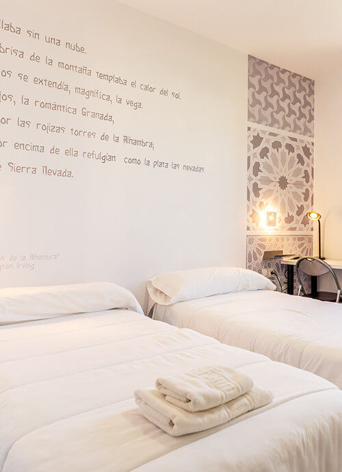 Interior de habitación en Albergue Inturjoven Granada, con edredones blancos, mesa de estudio e inscripciones árabes en las paredes.