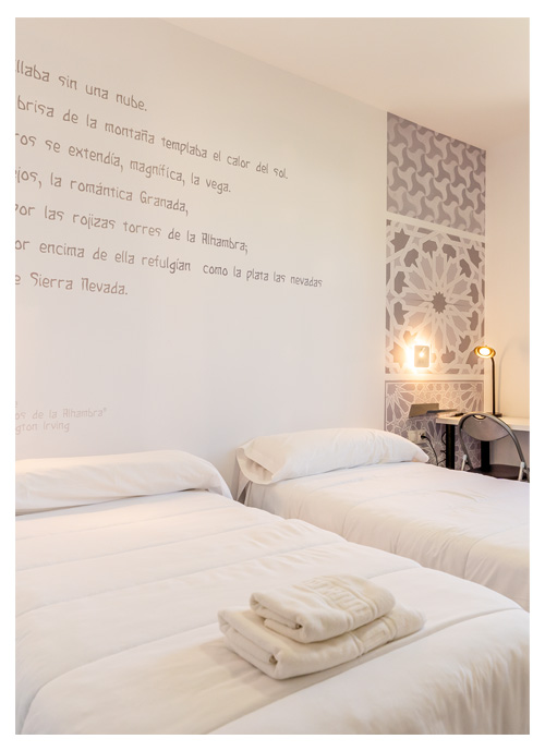 Interior de habitación en Albergue Inturjoven Granada, con edredones blancos, mesa de estudio e inscripciones árabes en las paredes.