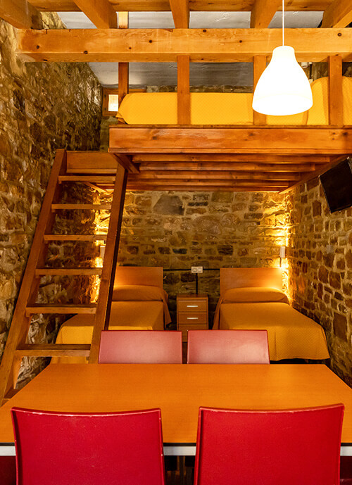 Interior de una de las cabañas de Inturjoven en Cortes de la Frontera, con 4 camas, dos alturas, paredes de piedra, cocina completa, baño y amplia mesa de comedor.