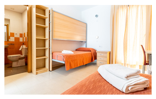 Habitación con 2 camas individuales, cama supletoria, baño completo propio, estanterías y mesa de estudio.